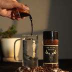 A premium glass jar of Bestia Brisk Original Instant Coffee is placed next to a clear mug filled with ice water, with a hand pouring rich black coffee concentrate into the glass. Fresh coffee beans are scattered at the base, and a blurred plant in the background adds a rustic, cozy touch. The Bestia Brisk logo, featuring a sleek black and gold design, is clearly visible, highlighting the brand's luxury appeal.