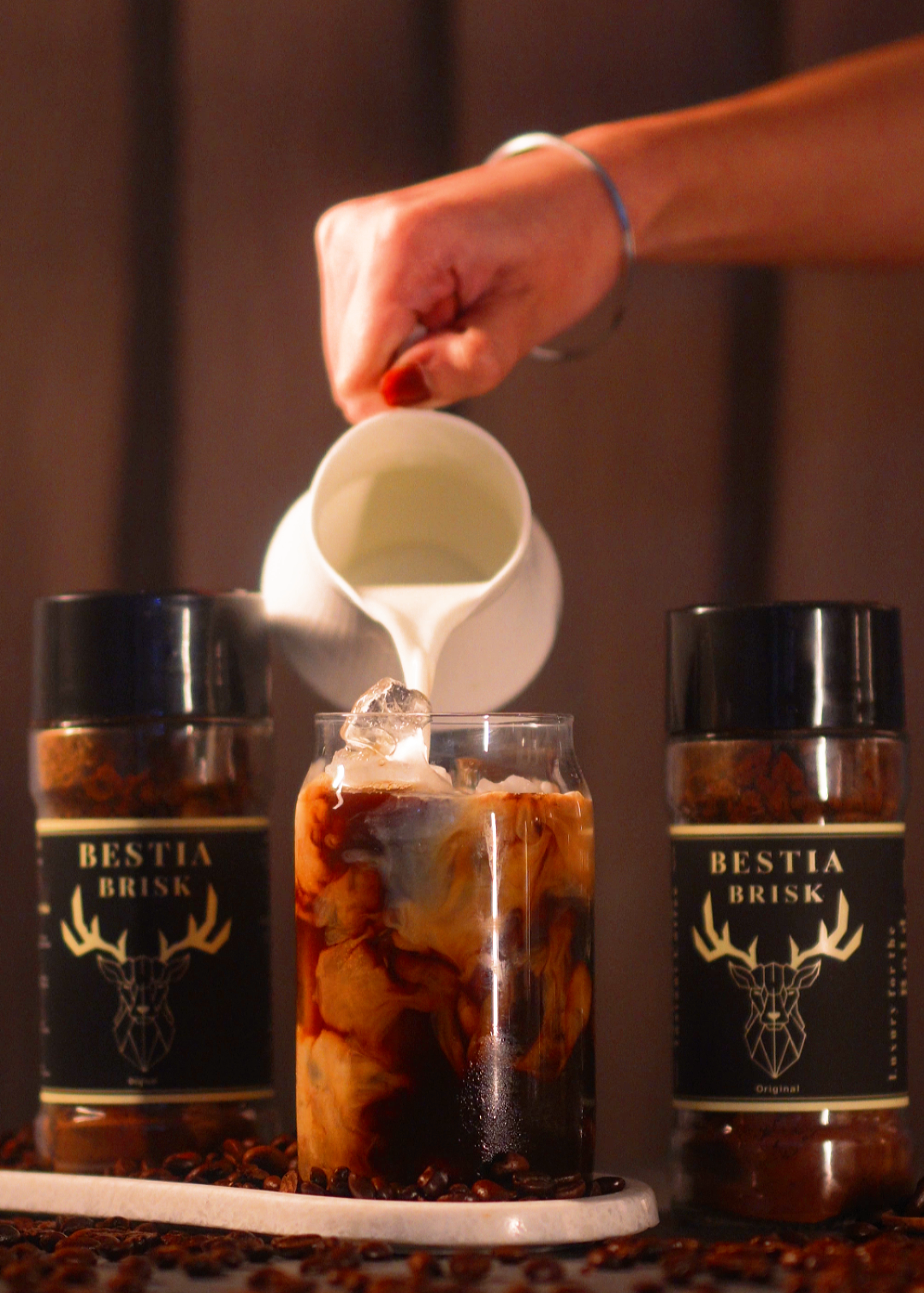 Person pouring cream into a glass of iced coffee with Bestia Brisk bottles in the background.